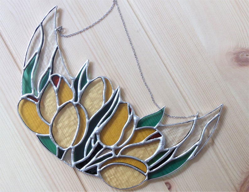 May include: Stained glass crescent moon with yellow tulip flowers, green leaves, and red accents. The glass is outlined with silver solder and hangs from a silver chain. The artwork is set against a light wood background.