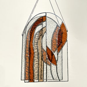 May include: Stained glass window panel with a clear arched top and a rectangular bottom. The design features vertical bands of orange, brown, and beige glass, with a leaf-like shape on the right side. A silver chain is attached for hanging.