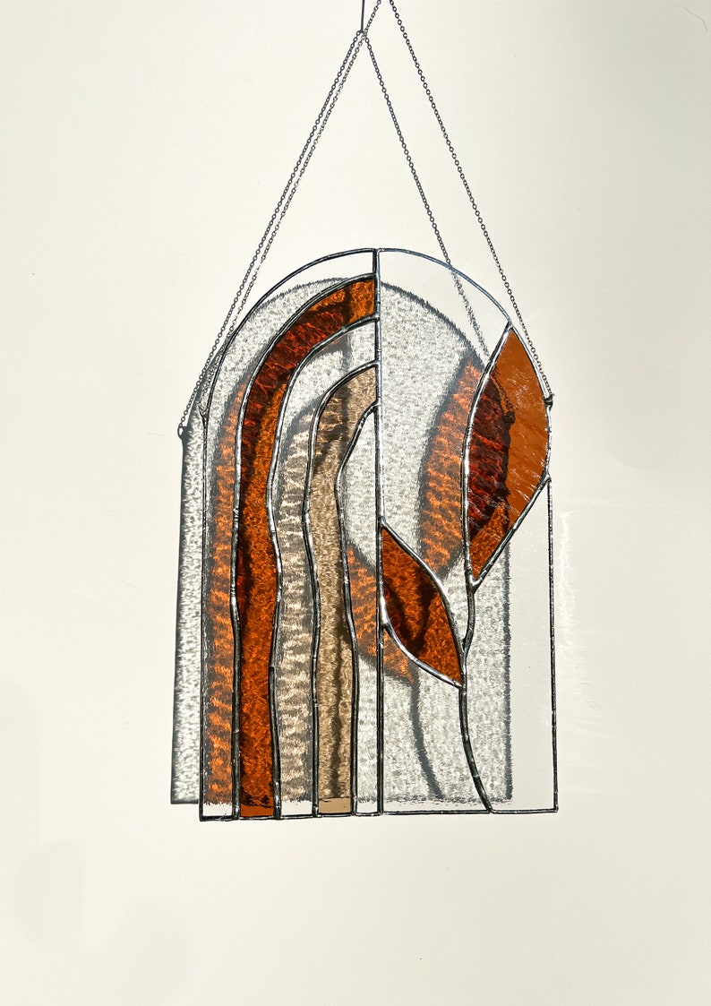 May include: Stained glass window panel with a clear arched top and a rectangular bottom. The design features vertical bands of orange, brown, and beige glass, with a leaf-like shape on the right side. A silver chain is attached for hanging.