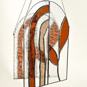 May include: Stained glass window panel with an arched top. The design features clear, orange, and tan glass sections, framed with black metal. The panel is suspended by a silver chain. The glass has a textured appearance.