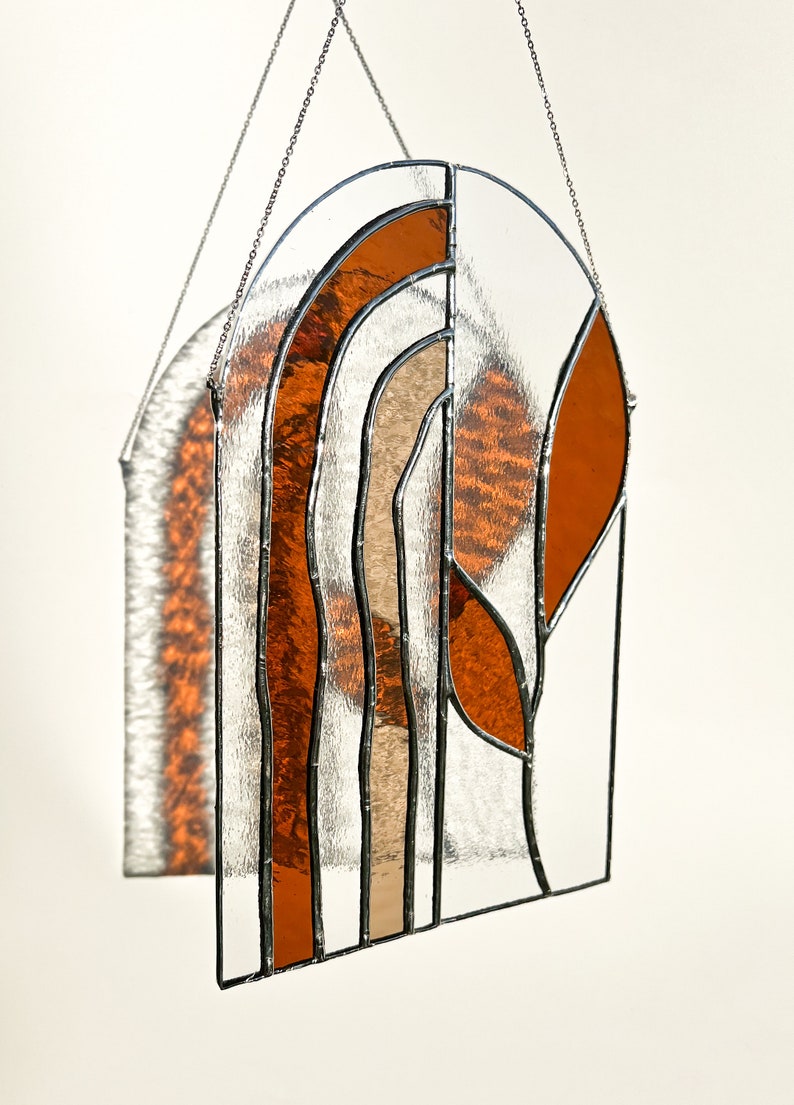 May include: Stained glass window panel with an arched top. The design features clear, orange, and tan glass sections, framed with black metal. The panel is suspended by a silver chain. The glass has a textured appearance.