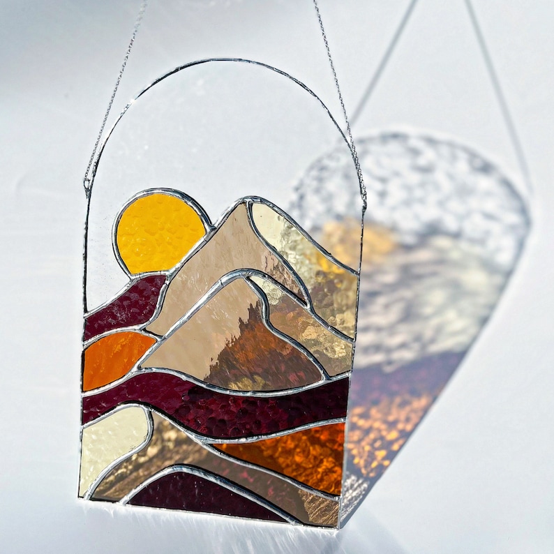 K&ouml;nnte beinhalten: Ein Buntglas-Sonnenf&auml;nger mit einer stilisierten Bergkette und einer gelben Sonne oben. Die Bergkette besteht aus verschiedenen farbigen Glasst&uuml;cken, darunter Braun, Rot, Orange und Wei&szlig;.