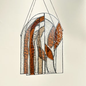 May include: Stained glass window panel with an arched top. The design features layered, textured glass in amber, brown, and clear. The panel is framed with silver metal and suspended by a chain.