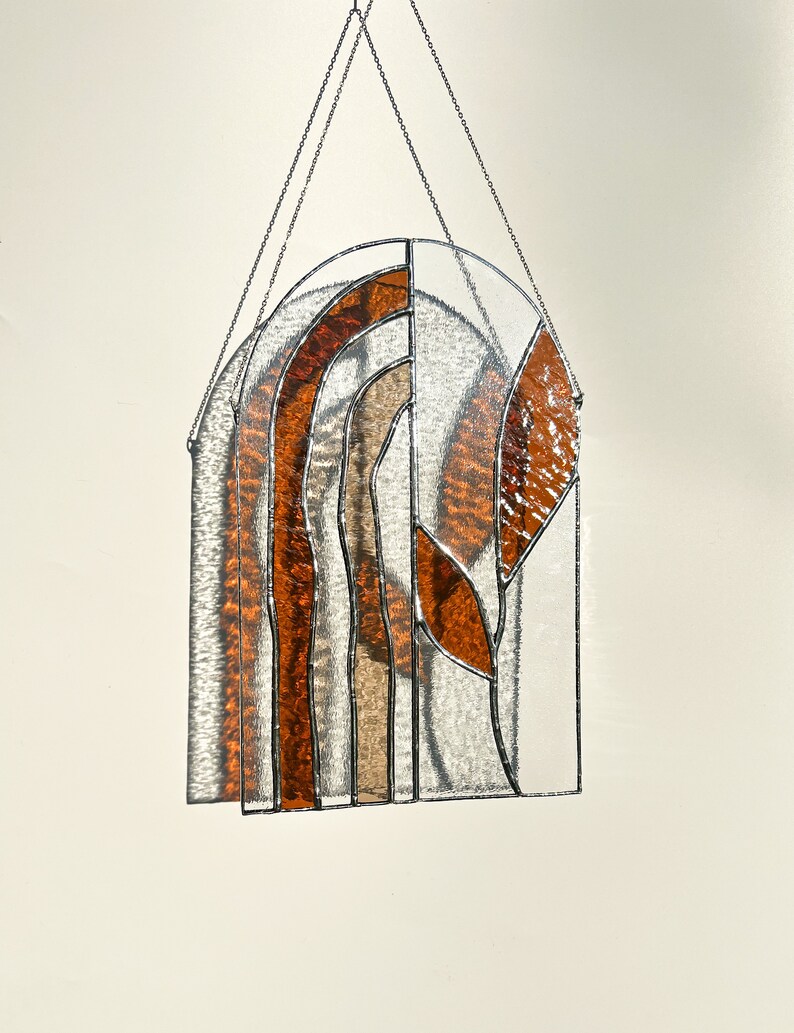 May include: Stained glass window panel with an arched top. The design features layered, textured glass in amber, brown, and clear. The panel is framed with silver metal and suspended by a chain.