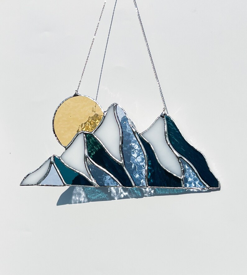 May include: A stained glass sun and mountain scene hanging from a silver chain. The sun is yellow and the mountains are blue, white, and green.