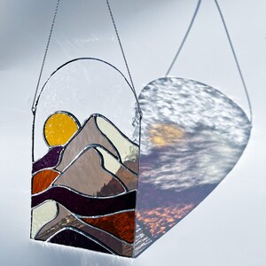 K&ouml;nnte beinhalten: Ein Buntglasfenster, das an einer Kette h&auml;ngt. Das Fenster zeigt einen Sonnenuntergang &uuml;ber einem Gebirge mit einer gelben Sonne, braunen Bergen und violetten und orangefarbenen Wellen.