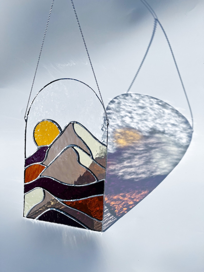 K&ouml;nnte beinhalten: Ein Buntglasfenster, das an einer Kette h&auml;ngt. Das Fenster zeigt einen Sonnenuntergang &uuml;ber einem Gebirge mit einer gelben Sonne, braunen Bergen und violetten und orangefarbenen Wellen.