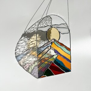 May include: A stained glass window hanging from a chain, depicting a colorful mountain range with a yellow sun and white clouds in the sky.