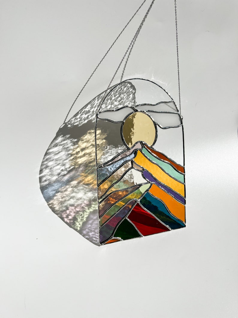 May include: A stained glass window hanging from a chain, depicting a colorful mountain range with a yellow sun and white clouds in the sky.