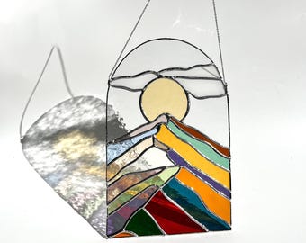 Può includere: Un acchiappasole in vetro colorato con parte superiore ad arco, un sole giallo pallido e cime montuose stratificate in tonalità vivaci di rosso, verde, blu, arancione e viola. L'oggetto è appeso a una sottile catena metallica su sfondo bianco.