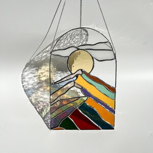 May include: A stained glass window hanging from a silver chain. The window depicts a stylized mountain range with a yellow sun and clouds in the sky. The mountains are made of different colored glass, including red, green, blue, purple, and yellow.