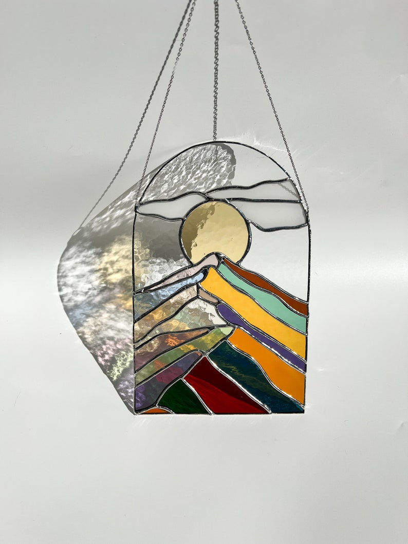 May include: A stained glass window hanging from a silver chain. The window depicts a stylized mountain range with a yellow sun and clouds in the sky. The mountains are made of different colored glass, including red, green, blue, purple, and yellow.