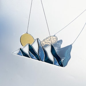May include: A stained glass sun and mountain scene hanging from a silver chain. The sun is yellow, the mountains are blue and white, and the background is a light blue.
