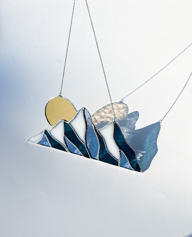 May include: A stained glass sun and mountain scene hanging from a silver chain. The sun is yellow, the mountains are blue and white, and the background is a light blue.