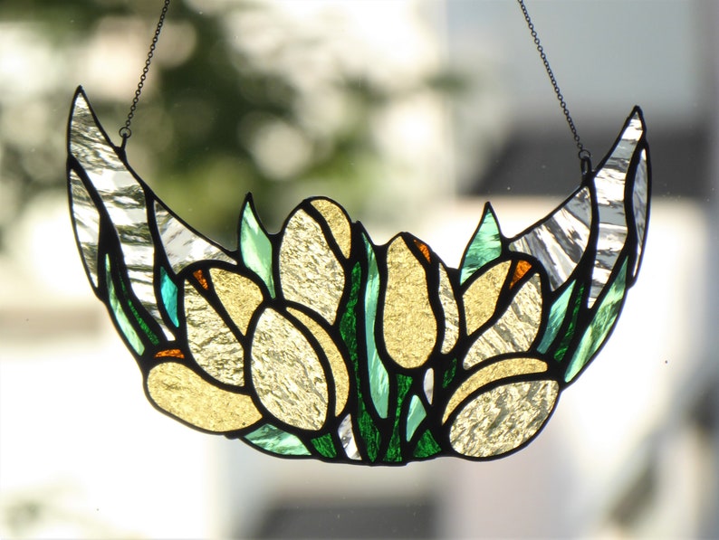 May include: Stained glass crescent moon with yellow tulip-like flowers and green leaves. The glass has a textured, iridescent quality, with black outlines and a chain for hanging. The design is reminiscent of Art Nouveau.