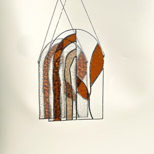 May include: Stained glass window hanging with an arched top. The design features a mix of clear, amber, and tan glass pieces, creating an abstract pattern. The piece is framed with black metal and suspended by a chain.
