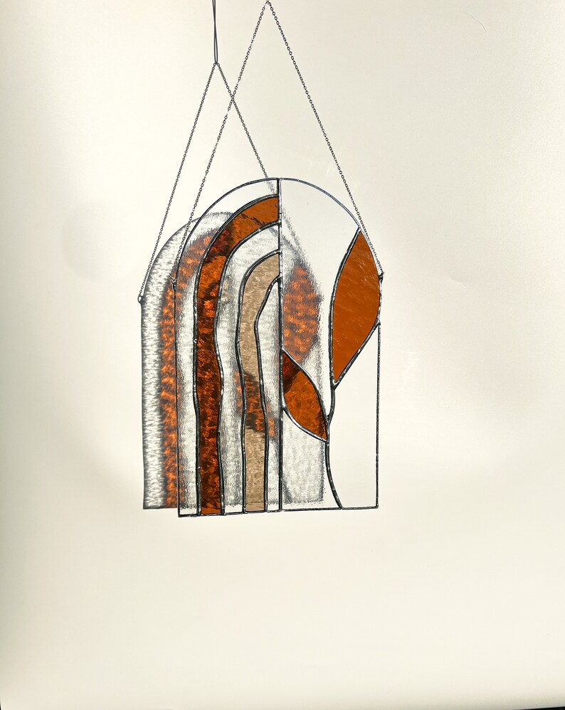 May include: Stained glass window hanging with an arched top. The design features a mix of clear, amber, and tan glass pieces, creating an abstract pattern. The piece is framed with black metal and suspended by a chain.
