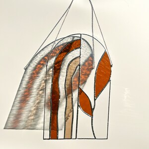 May include: Stained glass window hanging with a geometric design. The artwork features a series of curved and angular shapes in shades of orange, brown, and clear glass, outlined with black solder. A silver chain is attached for hanging.