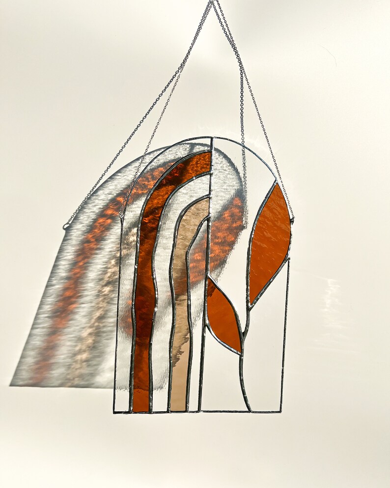 May include: Stained glass window hanging with a geometric design. The artwork features a series of curved and angular shapes in shades of orange, brown, and clear glass, outlined with black solder. A silver chain is attached for hanging.