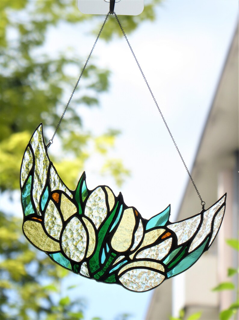 May include: A stained glass crescent moon suncatcher with a floral design. The piece features yellow, green, and clear textured glass, with a black chain for hanging. The design includes tulip-like shapes.