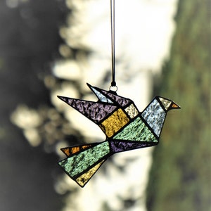 Pode incluir: Um ornamento de p&aacute;ssaro em vitral com um design geom&eacute;trico, suspenso por um cord&atilde;o preto. O p&aacute;ssaro apresenta um mosaico de pe&ccedil;as de vidro coloridas em tons de roxo, amarelo, verde e azul. O bico do p&aacute;ssaro &eacute; um pequeno tri&acirc;ngulo laranja.