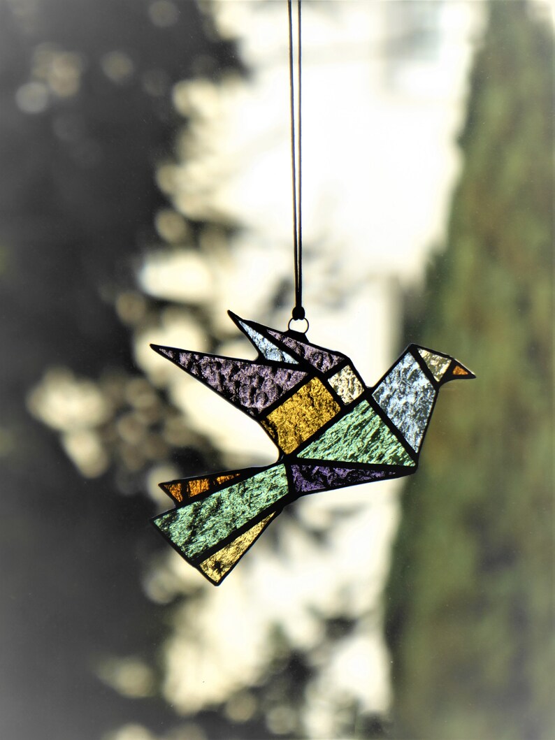 Pode incluir: Um ornamento de p&aacute;ssaro em vitral com um design geom&eacute;trico, suspenso por um cord&atilde;o preto. O p&aacute;ssaro apresenta um mosaico de pe&ccedil;as de vidro coloridas em tons de roxo, amarelo, verde e azul. O bico do p&aacute;ssaro &eacute; um pequeno tri&acirc;ngulo laranja.