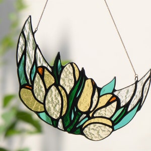 May include: Stained glass crescent moon with a floral design. The artwork features white, yellow, green, and teal glass pieces, outlined in black. A chain is attached for hanging. The design resembles tulips.