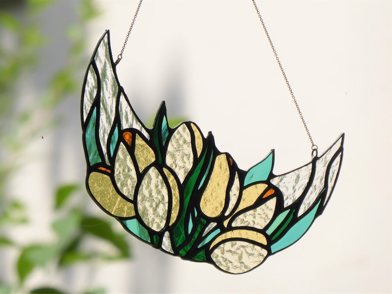 May include: Stained glass crescent moon with a floral design. The artwork features white, yellow, green, and teal glass pieces, outlined in black. A chain is attached for hanging. The design resembles tulips.