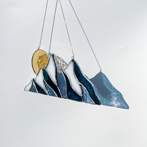 May include: A stained glass mountain range with a gold sun hanging from a silver chain. The mountains are blue and white with a textured glass effect.