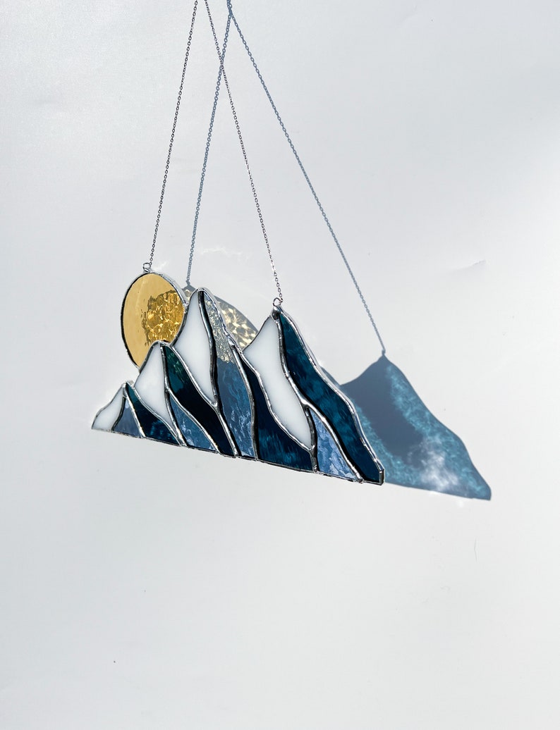 May include: A stained glass mountain range with a gold sun hanging from a silver chain. The mountains are blue and white with a textured glass effect.