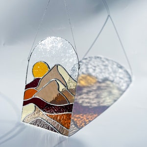 K&ouml;nnte beinhalten: Ein Buntglasfenster, das an einer Kette h&auml;ngt. Das Fenster zeigt einen Sonnenuntergang &uuml;ber einem Gebirge mit einer gelben Sonne, braunen Bergen und roten, orangefarbenen und gelben Akzenten.