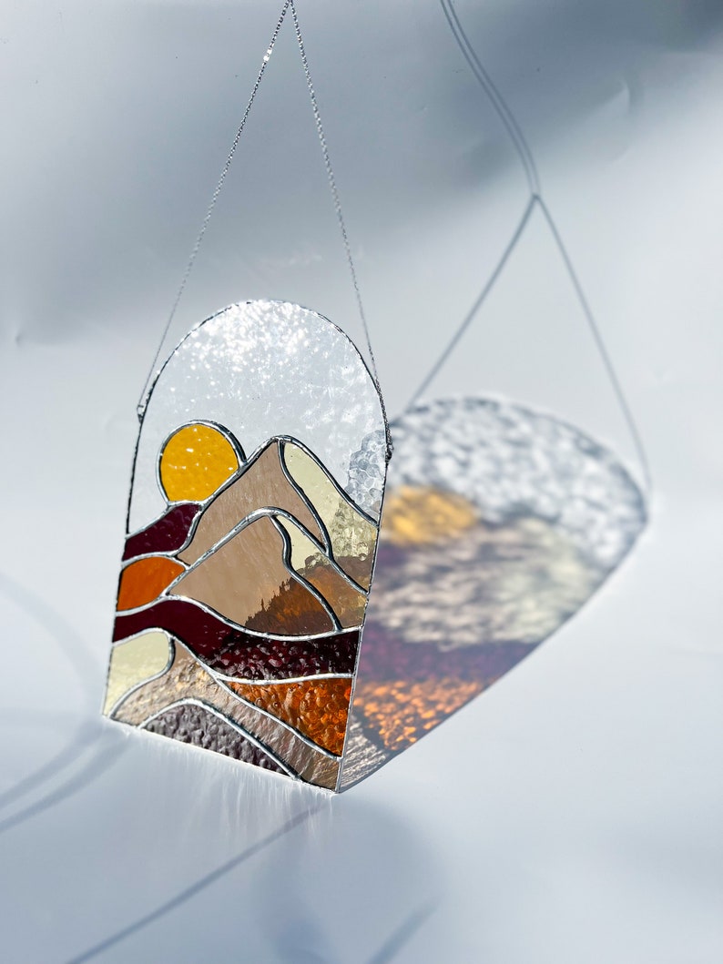 K&ouml;nnte beinhalten: Ein Buntglasfenster, das an einer Kette h&auml;ngt. Das Fenster zeigt einen Sonnenuntergang &uuml;ber einem Gebirge mit einer gelben Sonne, braunen Bergen und roten, orangefarbenen und gelben Akzenten.