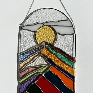 May include: A stained glass window hanging with a chain. The window depicts a stylized mountain range with a yellow sun and a white cloud in the sky. The mountain range is made of colorful glass pieces in shades of red, orange, yellow, green, blue, purple, and pink.