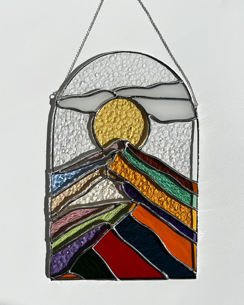 May include: A stained glass window hanging with a chain. The window depicts a stylized mountain range with a yellow sun and a white cloud in the sky. The mountain range is made of colorful glass pieces in shades of red, orange, yellow, green, blue, purple, and pink.
