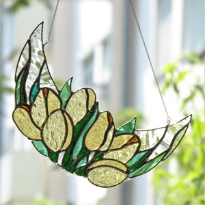 May include: A stained glass crescent moon with yellow tulip-like flowers and green leaves. The artwork is suspended by a silver chain. The glass has clear and textured sections, and the piece is set against a blurred background of greenery.