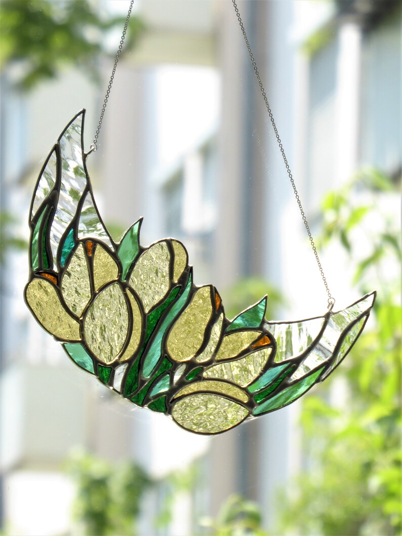 May include: A stained glass crescent moon with yellow tulip-like flowers and green leaves. The artwork is suspended by a silver chain. The glass has clear and textured sections, and the piece is set against a blurred background of greenery.
