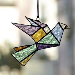 Pode incluir: Um apanhador de sol de p&aacute;ssaro origami em vitral. O p&aacute;ssaro &eacute; composto por formas geom&eacute;tricas em tons de roxo, dourado, verde e azul. Ele &eacute; pendurado em um cord&atilde;o preto, ideal para decora&ccedil;&atilde;o de janelas ou paredes.