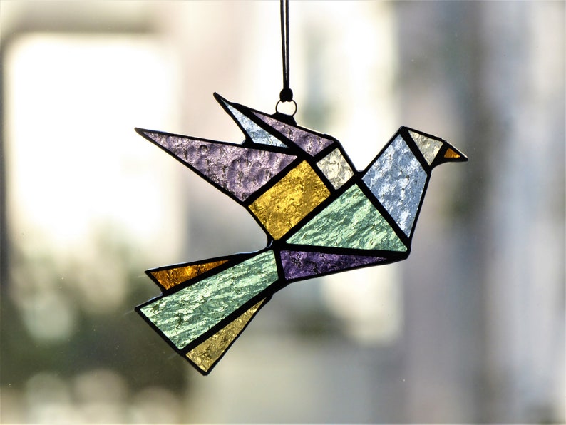 Pode incluir: Um apanhador de sol de p&aacute;ssaro origami em vitral. O p&aacute;ssaro &eacute; composto por formas geom&eacute;tricas em tons de roxo, dourado, verde e azul. Ele &eacute; pendurado em um cord&atilde;o preto, ideal para decora&ccedil;&atilde;o de janelas ou paredes.