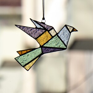 Pode incluir: Uma pomba de origami em vitral em voo, com formas geom&eacute;tricas em tons de roxo, dourado, verde e azul. O p&aacute;ssaro &eacute; delineado em preto e suspenso por um cord&atilde;o preto, criando um apanhador de sol decorativo.
