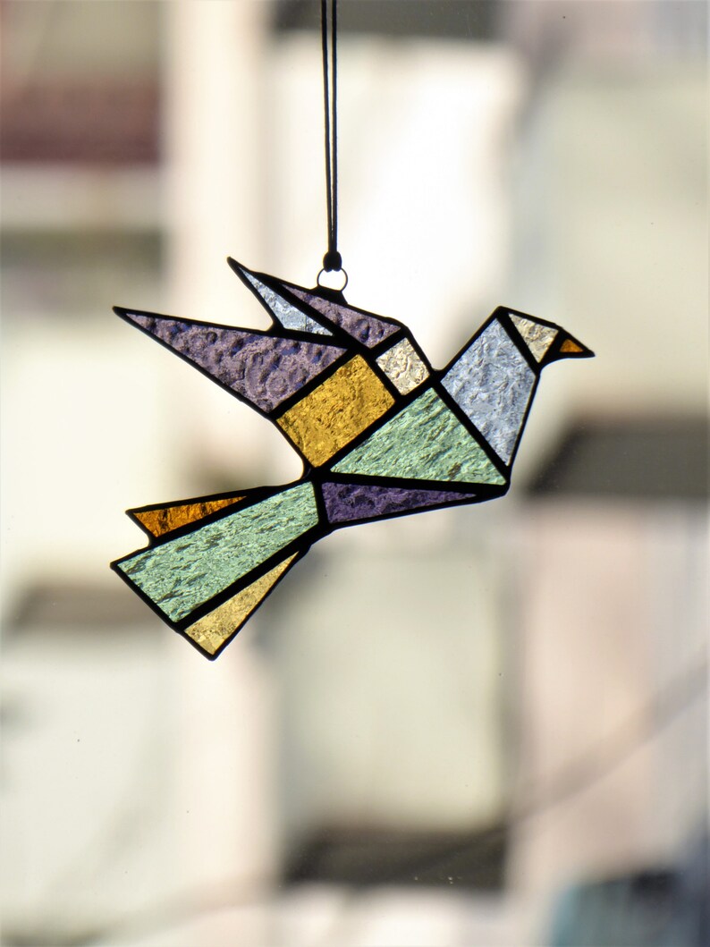 Pode incluir: Uma pomba de origami em vitral em voo, com formas geom&eacute;tricas em tons de roxo, dourado, verde e azul. O p&aacute;ssaro &eacute; delineado em preto e suspenso por um cord&atilde;o preto, criando um apanhador de sol decorativo.