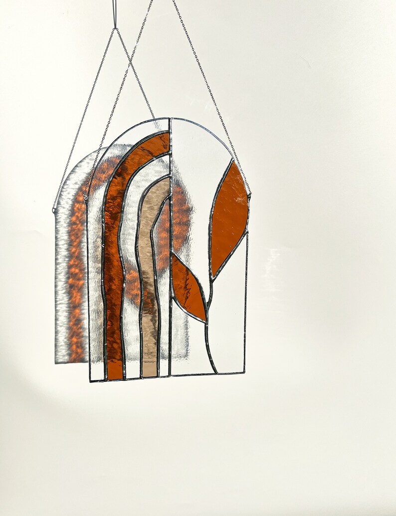 May include: Stained glass window panel with a geometric design. The panel features a half-circle shape with clear, brown, and orange glass pieces. The panel is suspended by two silver chains, creating a decorative and artistic piece.