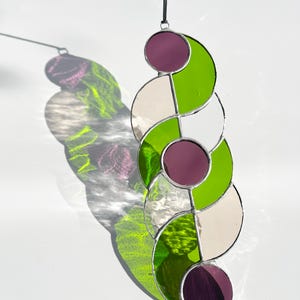 Puede incluir: Un atrapasoles de vidriera con un dise&ntilde;o geom&eacute;trico. Presenta semic&iacute;rculos verdes, blancos y morados alternados, unidos por soldadura plateada. Cuelga de un cord&oacute;n negro, proyectando una sombra colorida.