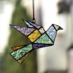Pode incluir: Um ornamento de p&aacute;ssaro origami em vitral. O p&aacute;ssaro apresenta um design geom&eacute;trico com v&aacute;rias cores, incluindo roxo, dourado, verde e azul. &Eacute; suspenso por um fio preto, adequado para pendurar. As asas do p&aacute;ssaro est&atilde;o abertas.