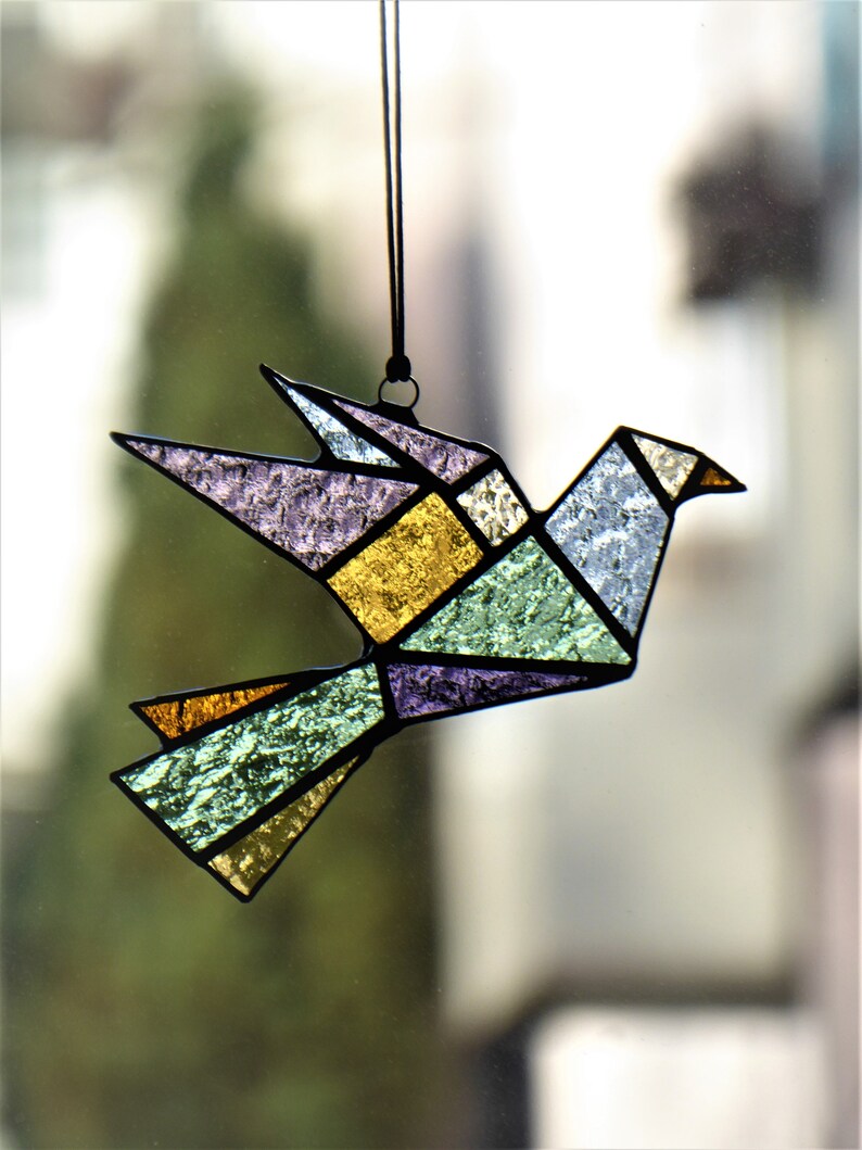 Pode incluir: Um ornamento de p&aacute;ssaro origami em vitral. O p&aacute;ssaro apresenta um design geom&eacute;trico com v&aacute;rias cores, incluindo roxo, dourado, verde e azul. &Eacute; suspenso por um fio preto, adequado para pendurar. As asas do p&aacute;ssaro est&atilde;o abertas.