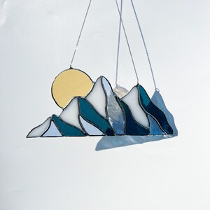 May include: A stained glass sun and mountain scene hanging from a silver chain. The sun is yellow and the mountains are blue and white.