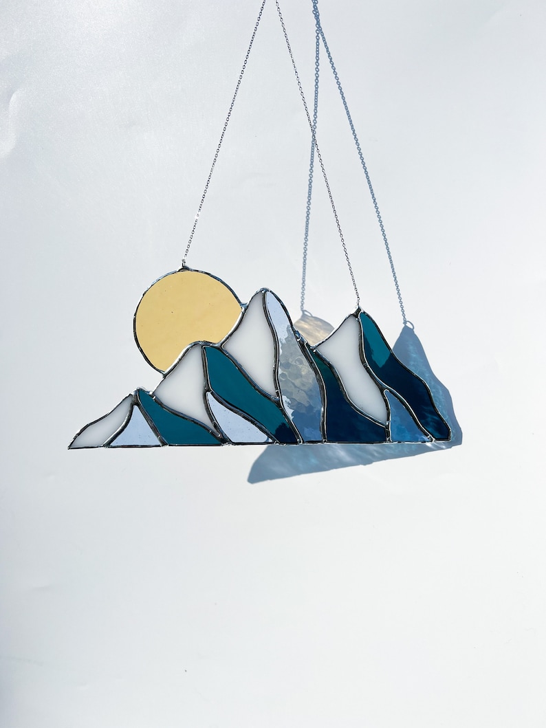 May include: A stained glass sun and mountain scene hanging from a silver chain. The sun is yellow and the mountains are blue and white.