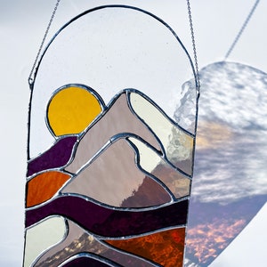 K&ouml;nnte beinhalten: Ein Buntglasfenster, das an einer Kette h&auml;ngt. Das Fenster zeigt einen Sonnenuntergang &uuml;ber einem Gebirge mit einer gelben Sonne, violetten, orangefarbenen und wei&szlig;en Bergen und einem wei&szlig;en Himmel.