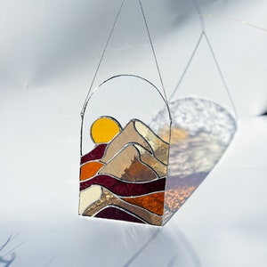 K&ouml;nnte beinhalten: Eine Sonne und eine Berglandschaft aus Buntglas, die an einer silbernen Kette h&auml;ngt. Die Sonne ist gelb und orange, die Berge sind braun und beige, und der Himmel ist dunkelrot.