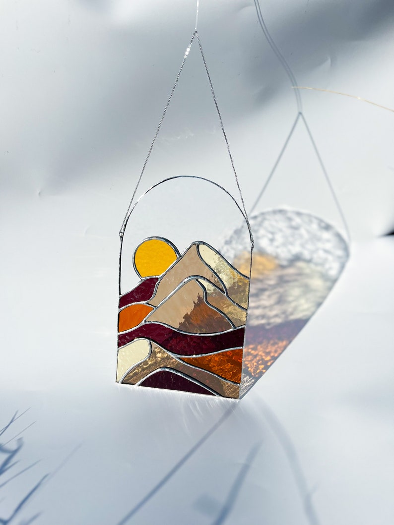 K&ouml;nnte beinhalten: Eine Sonne und eine Berglandschaft aus Buntglas, die an einer silbernen Kette h&auml;ngt. Die Sonne ist gelb und orange, die Berge sind braun und beige, und der Himmel ist dunkelrot.