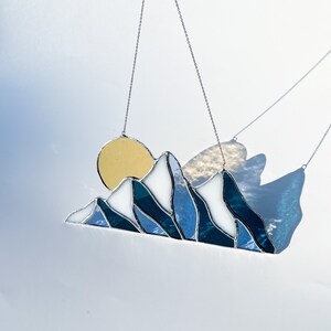 May include: A stained glass sun and mountain scene hanging from a silver chain. The sun is yellow, the mountains are white and blue, and the background is a light blue.